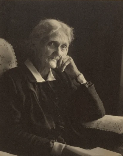 Black and white photo of an elderly woman leaning on her hand, sitting in an old chair and looking at the camera