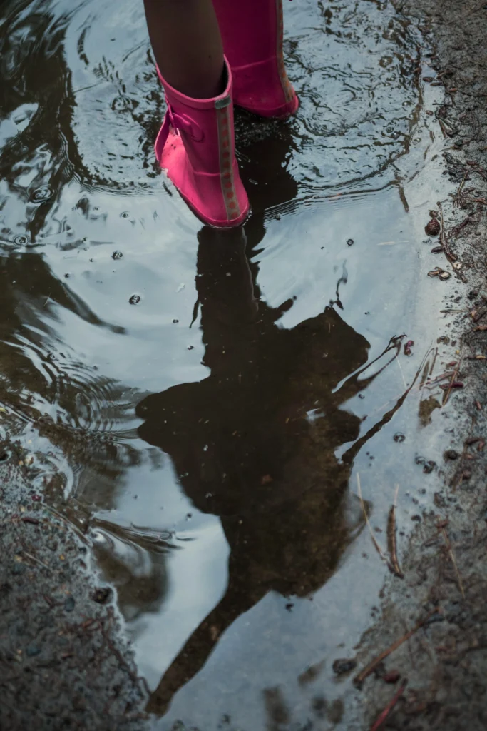 Child’s pink boots splash in a puddle, reflection shimmering below.